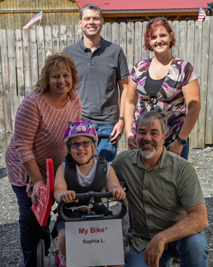 A young girl smiles while seated on an adaptive bicycle, surrounded by her parents and members of LHORBA's board.
